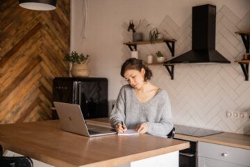 vrouw zit in aan een keukentafel achter haar laptop en schrijft in een notitieboek als afbeelding bij een blog over valkuil pensioen